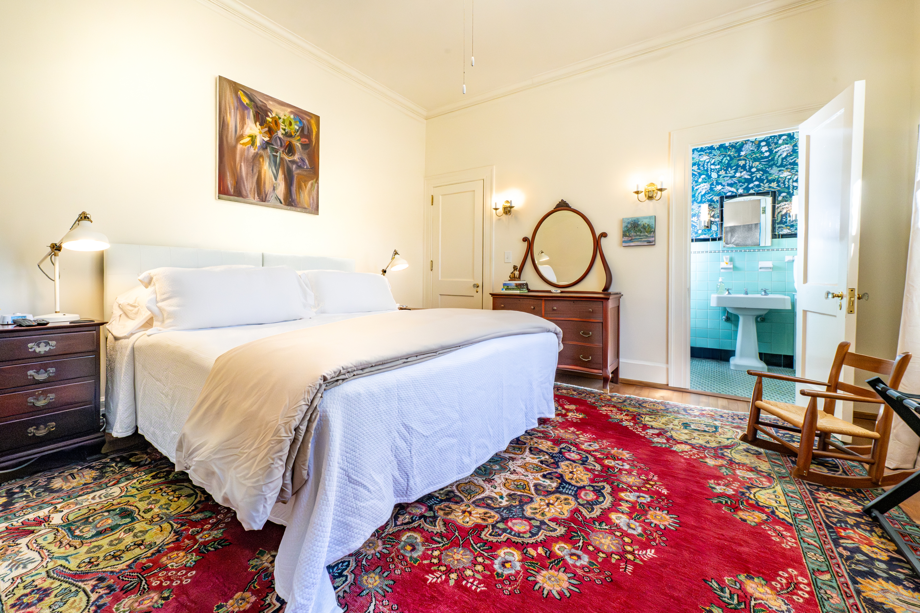 Light-filled bedroom with a plush white bed, large red rug, and antique dresser. An open door reveals a charming teal-tiled en suite bathroom.