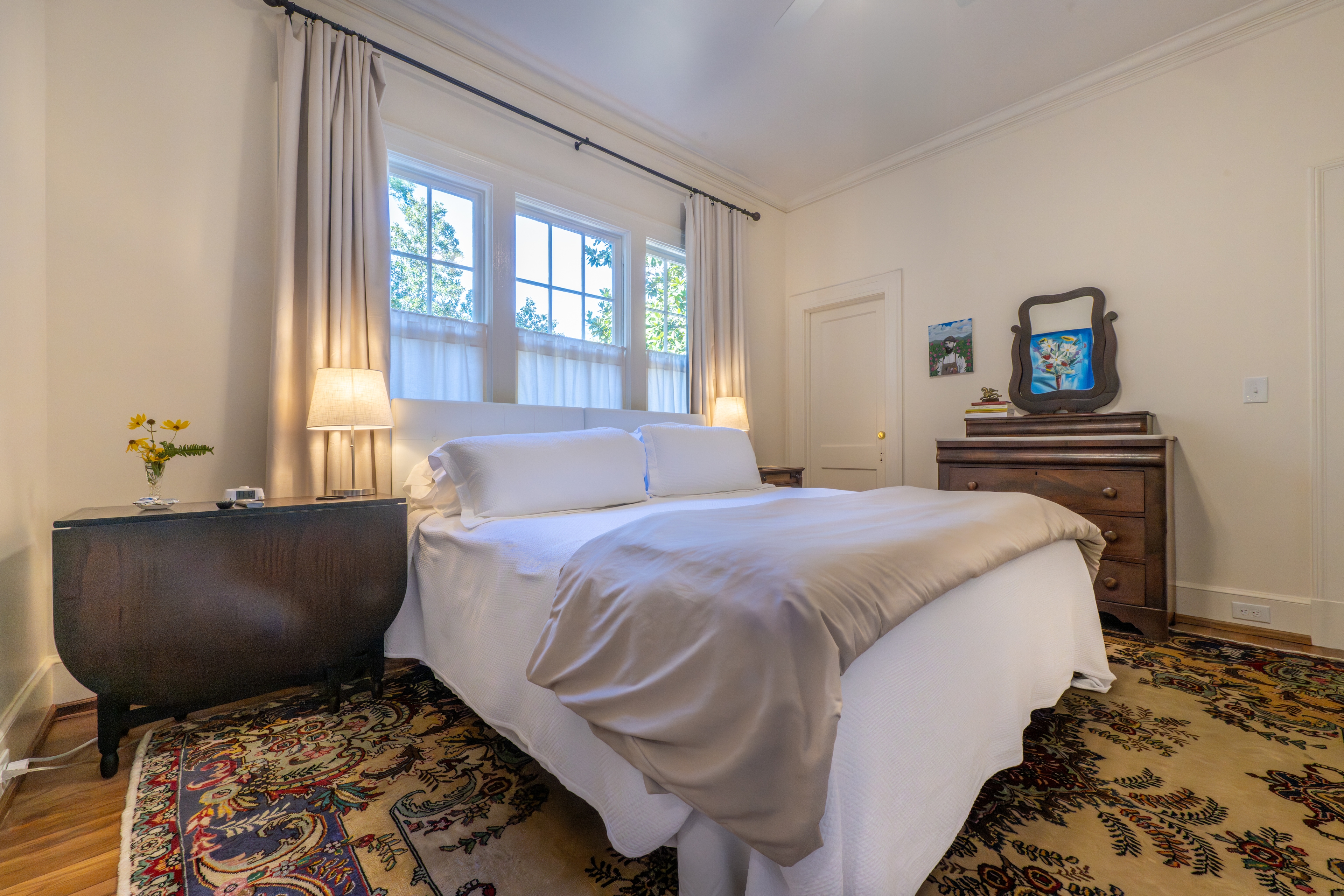 Sunlit bedroom with a white bed, beige patterned rug, and antique drop-leaf table. Three windows with curtains fill the room with natural light.