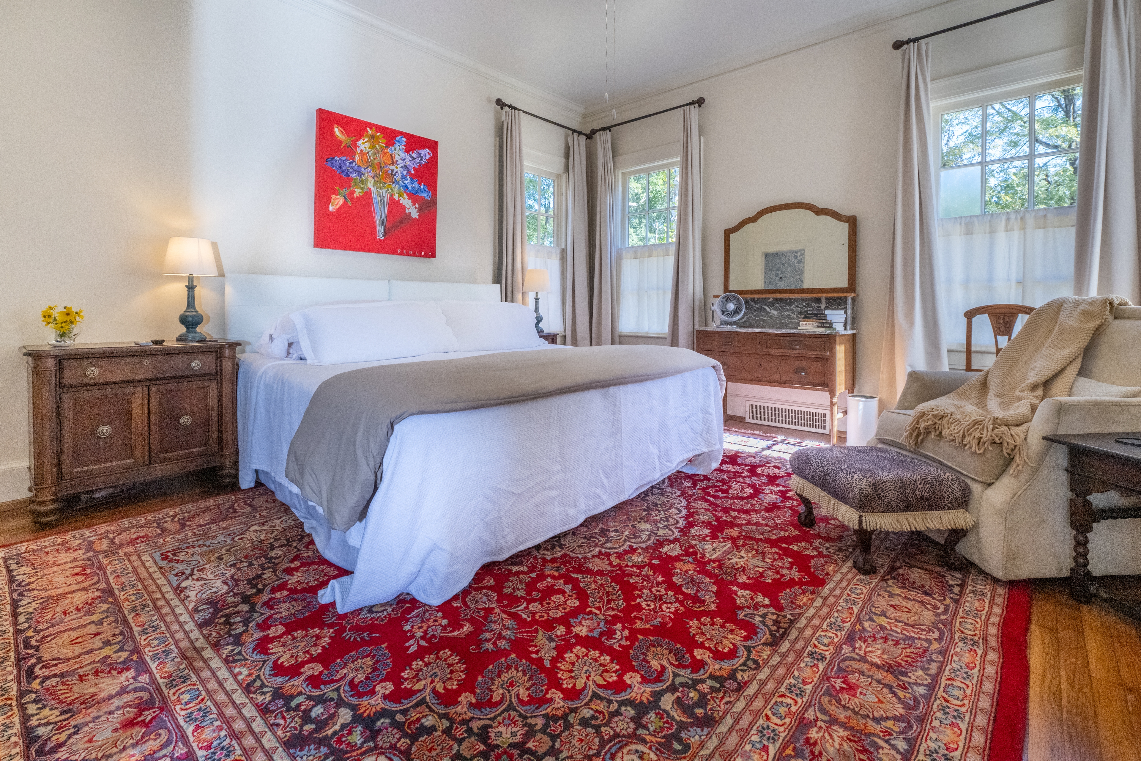 Light-filled bedroom with a plush white bed, large red rug, and antique dresser. An open door reveals a charming teal-tiled en suite bathroom.