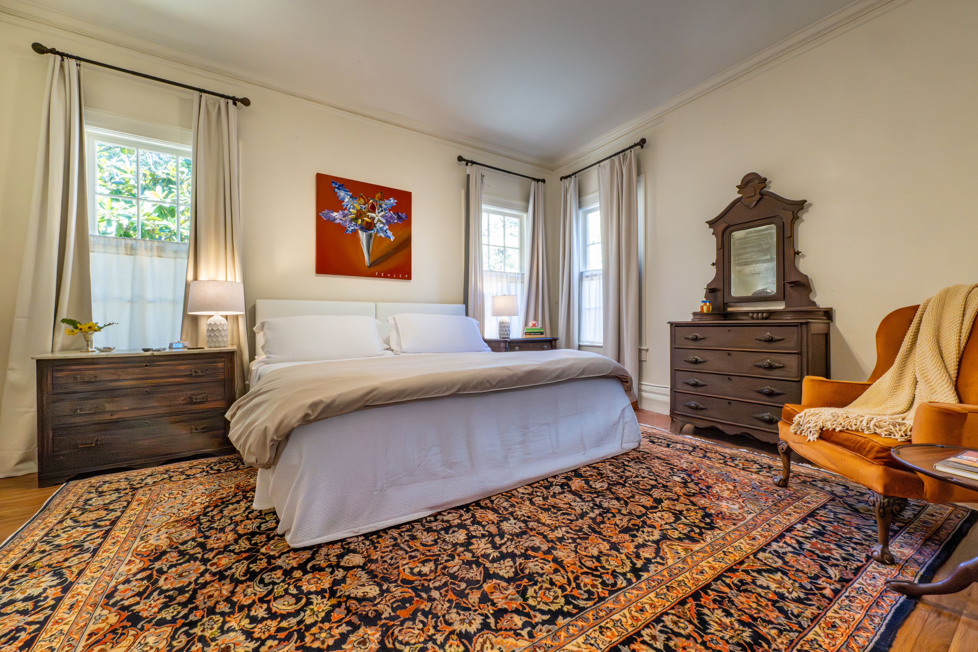 Warm bedroom featuring a large ornate rug, antique dresser with mirror, and a cozy orange velvet armchair. Natural light brightens the historic room.