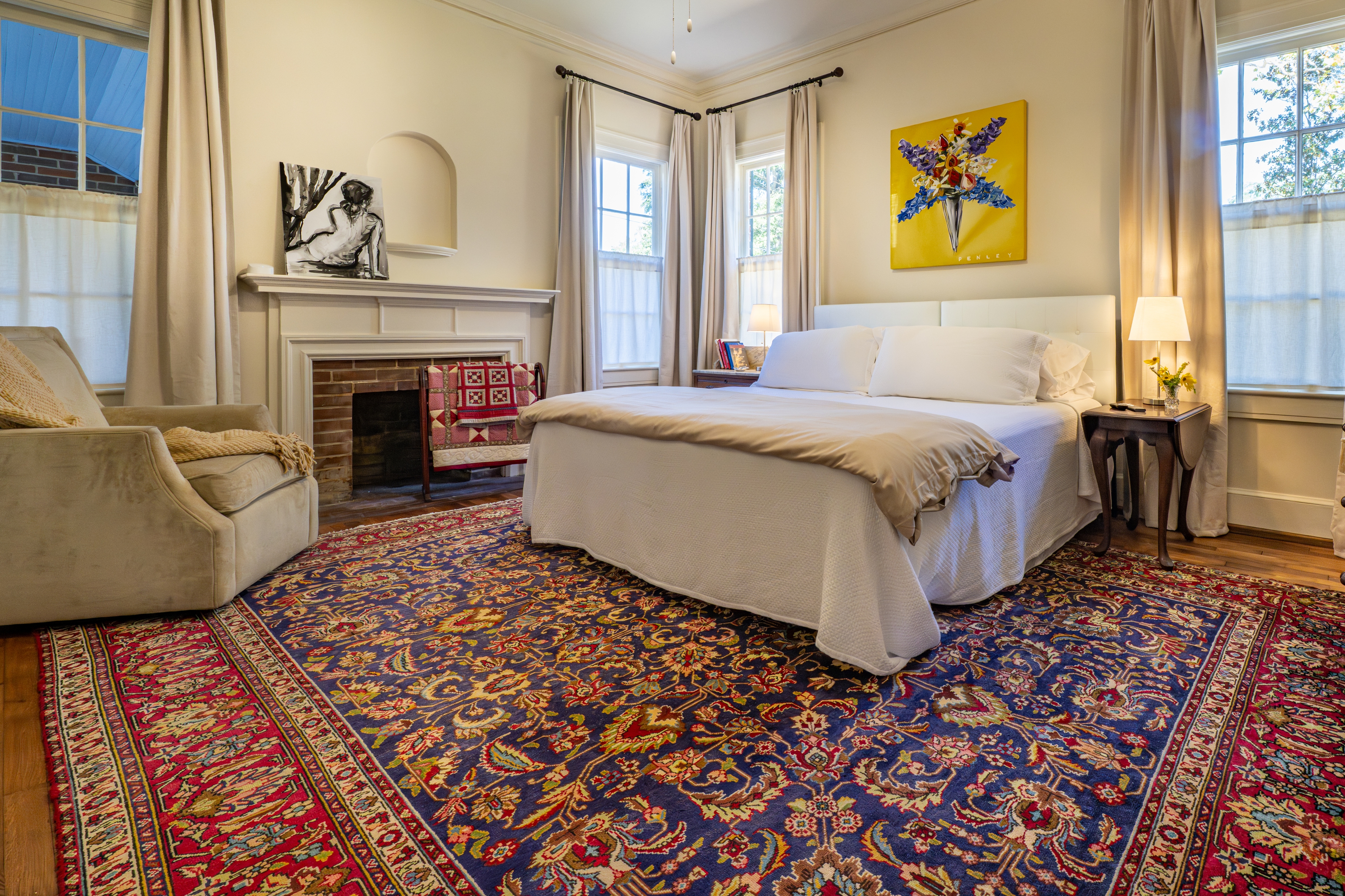 Spacious bedroom featuring a fireplace, vibrant patterned rug, and plush white bed. Includes a cozy armchair and bright yellow art piece.