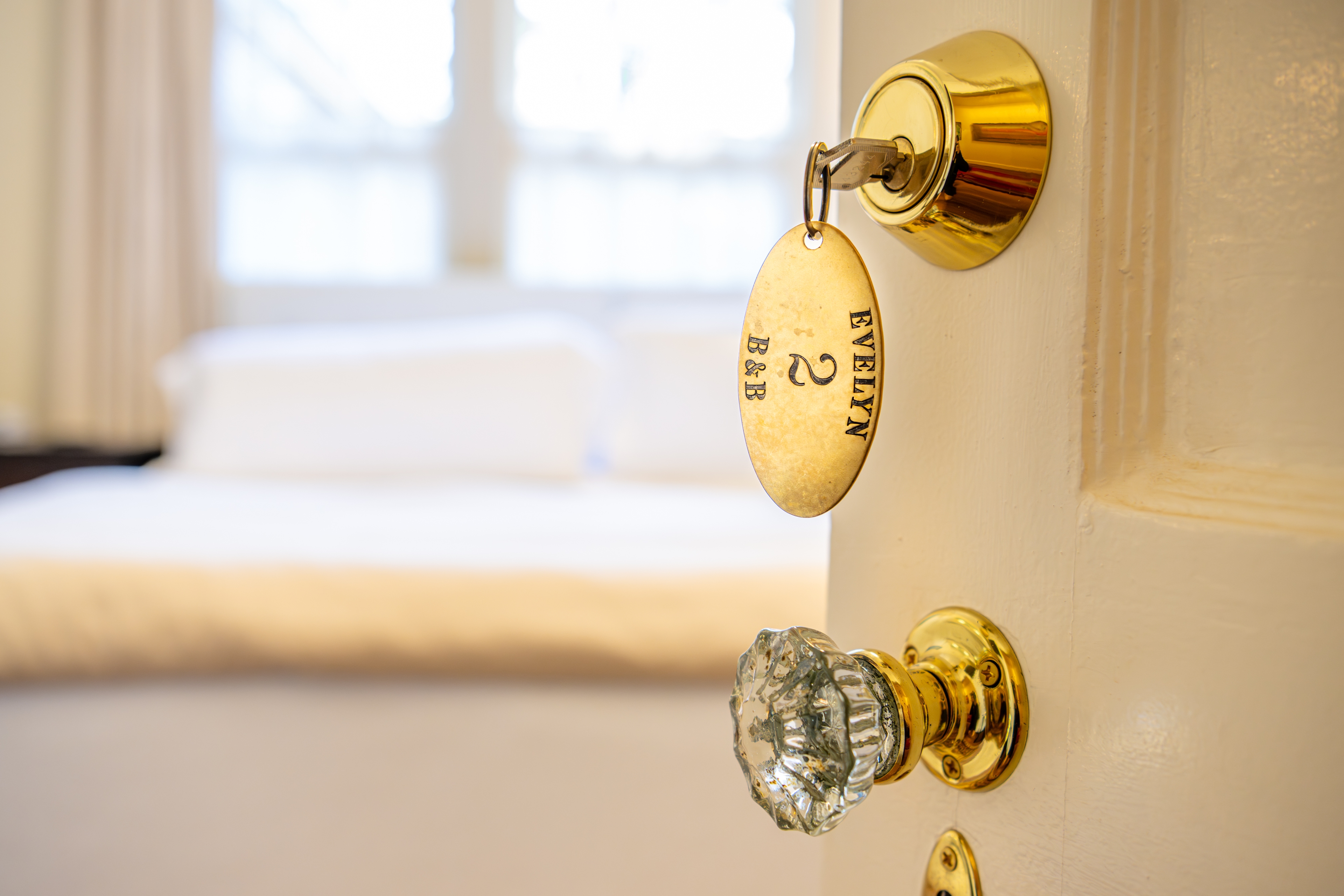 Close-up of a brass key with an "Evelyn" tag in a white door. A soft, sunlit bedroom with fresh linens is visible in the blurred background.