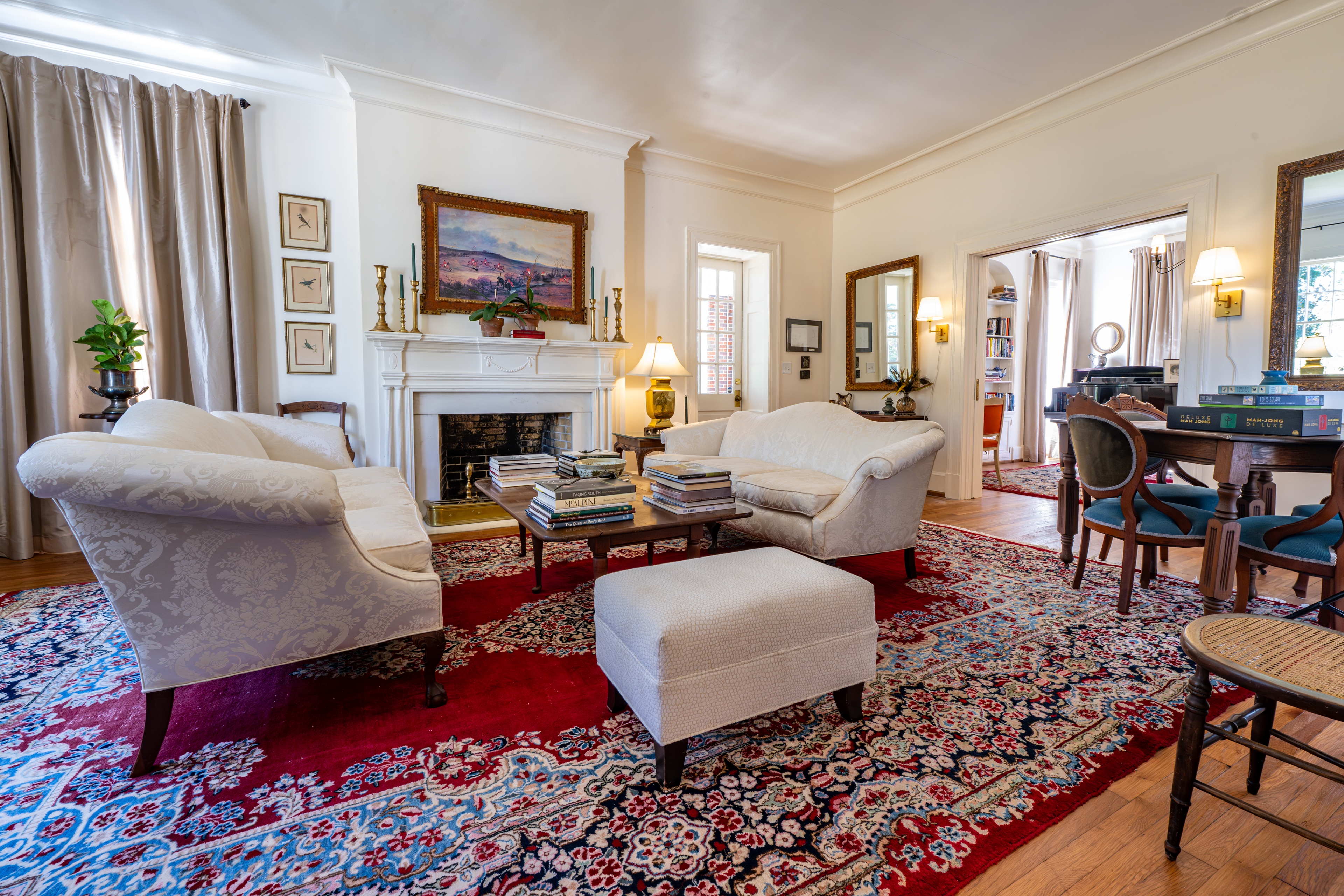 Elegant living room featuring white sofas, a rich red rug, and a classic fireplace. Sunlight illuminates the antique decor and warm hardwood floors.