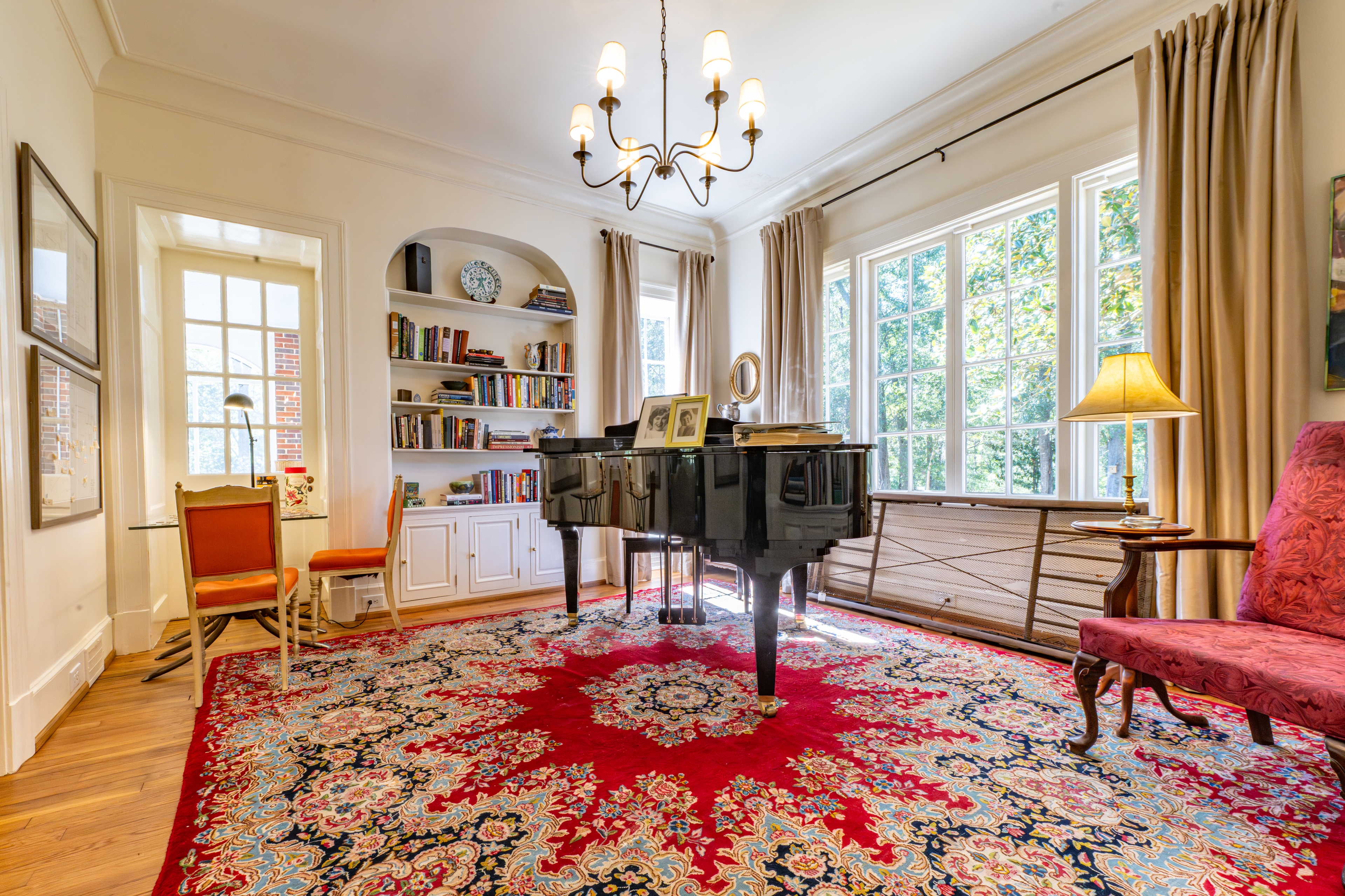 Elegant music room featuring a grand piano, built-in library shelves, and a vibrant red rug. Large windows fill the space with natural light.