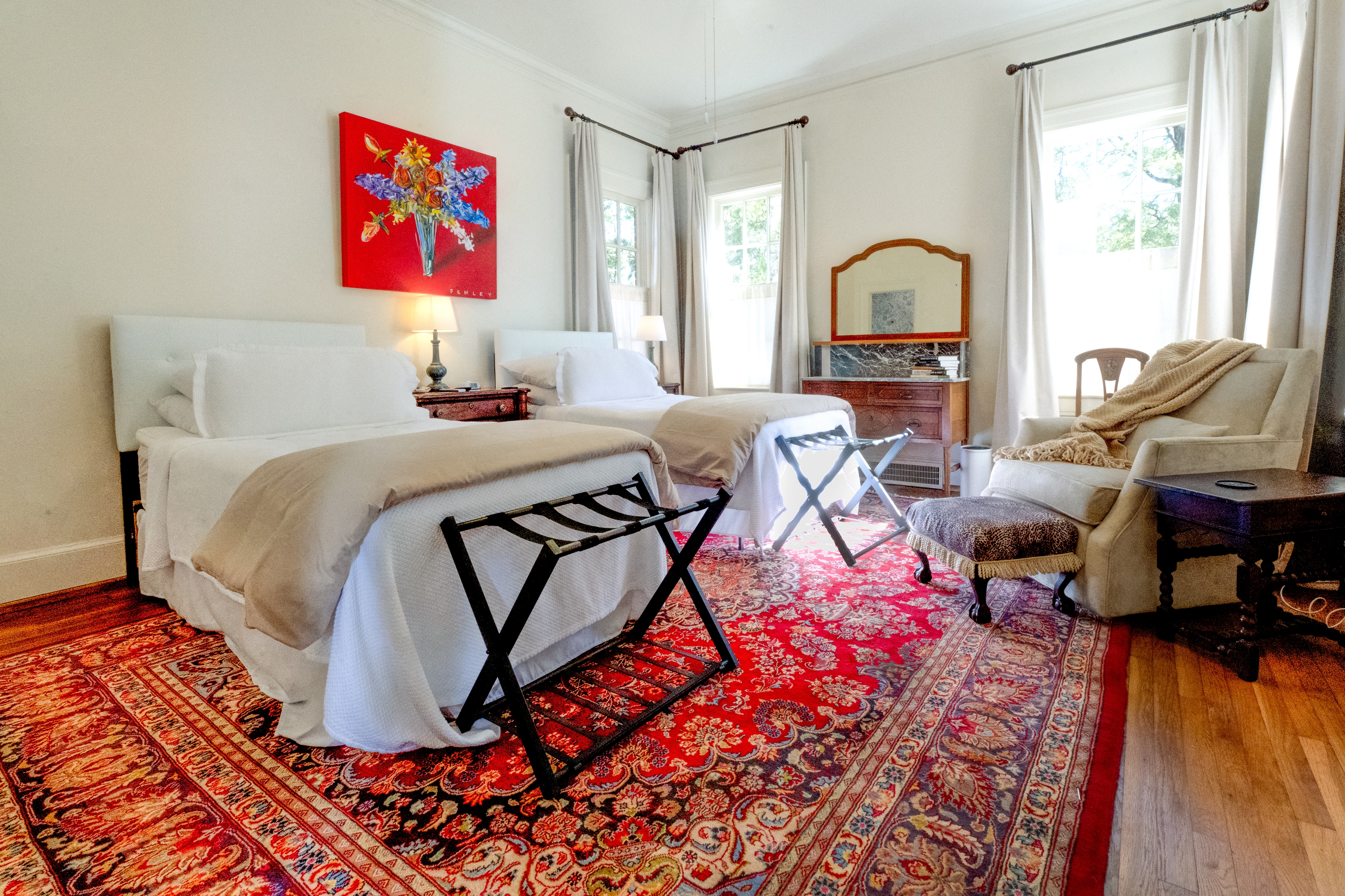 Twin bedroom featuring a vibrant red rug and bold red art. Includes a white armchair, luggage racks, and large windows.