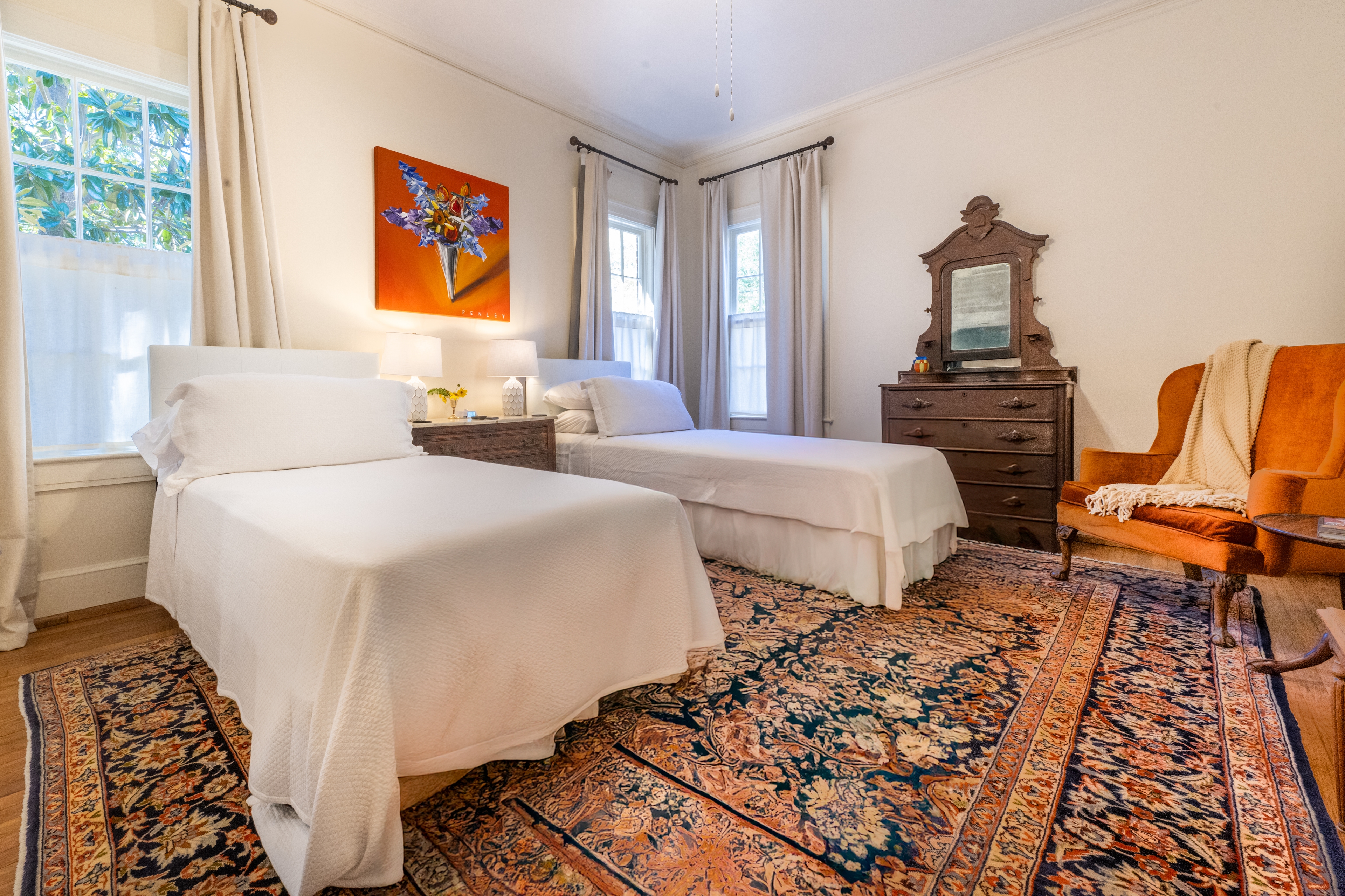 Charming bedroom featuring two twin beds, a blue oriental rug, and a plush orange armchair. Antique furniture and bright art complete the look.