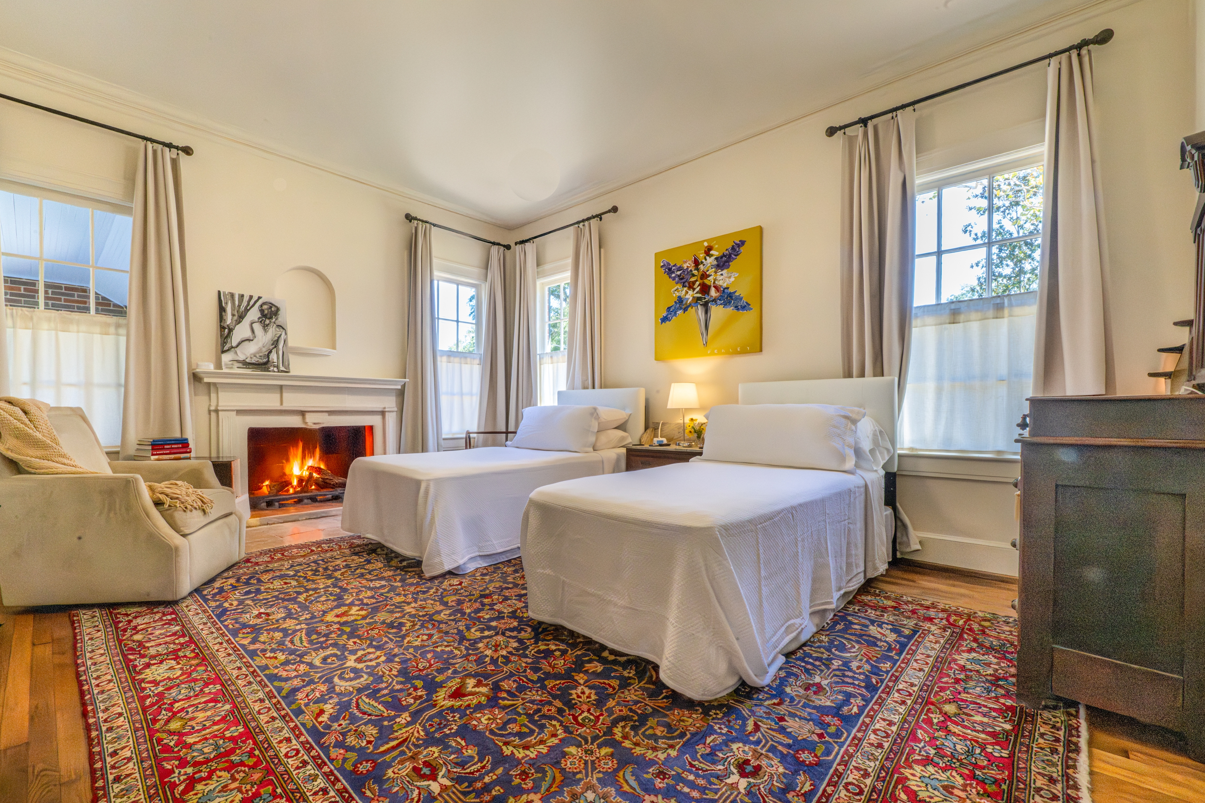 Cozy twin bedroom featuring a warm fireplace, blue patterned rug, and bright yellow art. Includes a plush armchair and antique dresser.
