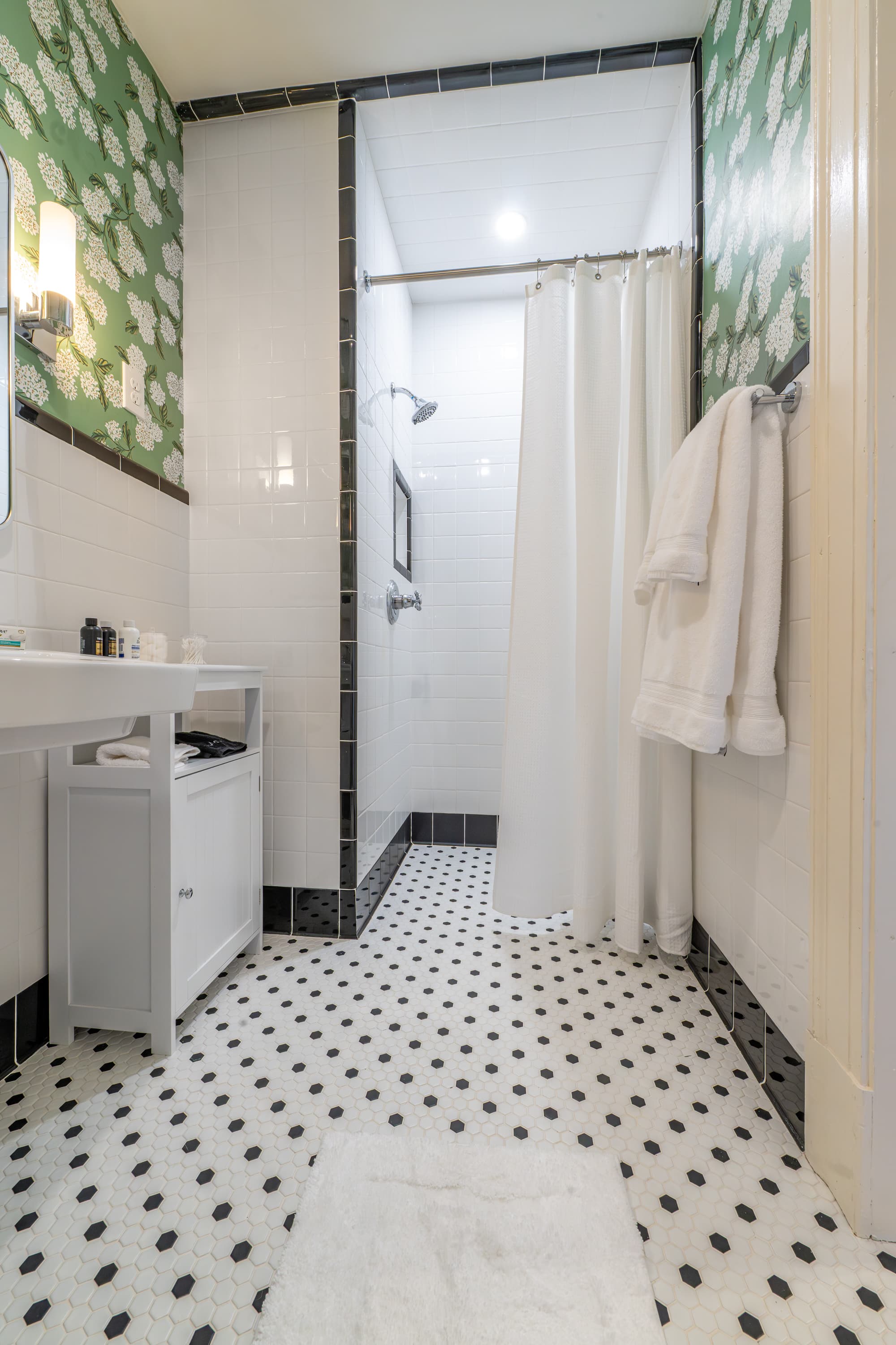 Accessible bathroom with a roll-in shower, green patterned wallpaper, and classic black-and-white hex tiles, combining safety with vintage style.