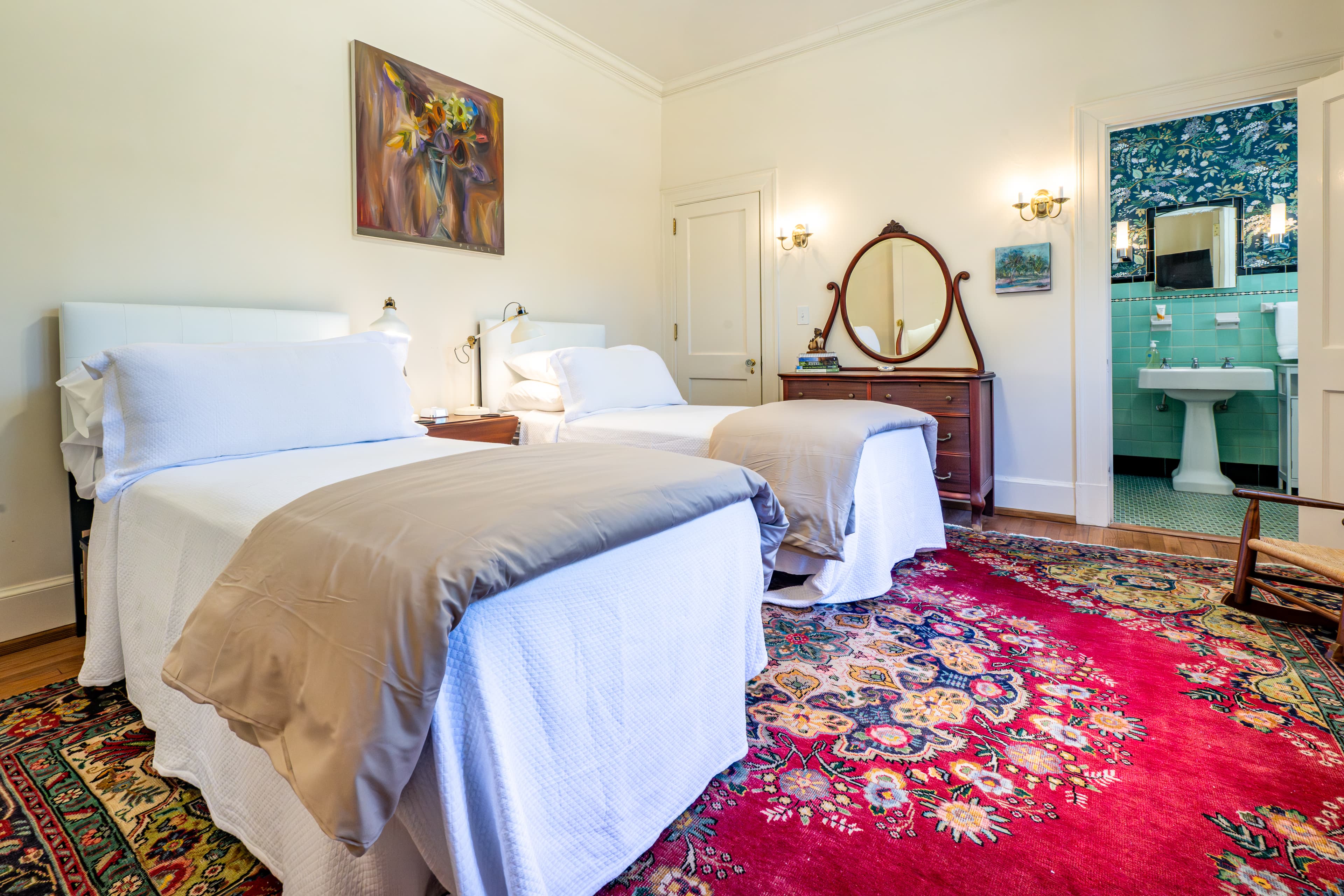 Spacious bedroom with a white bed, red rug, and antique dresser. An open door reveals a bathroom with teal walls and a pedestal sink.