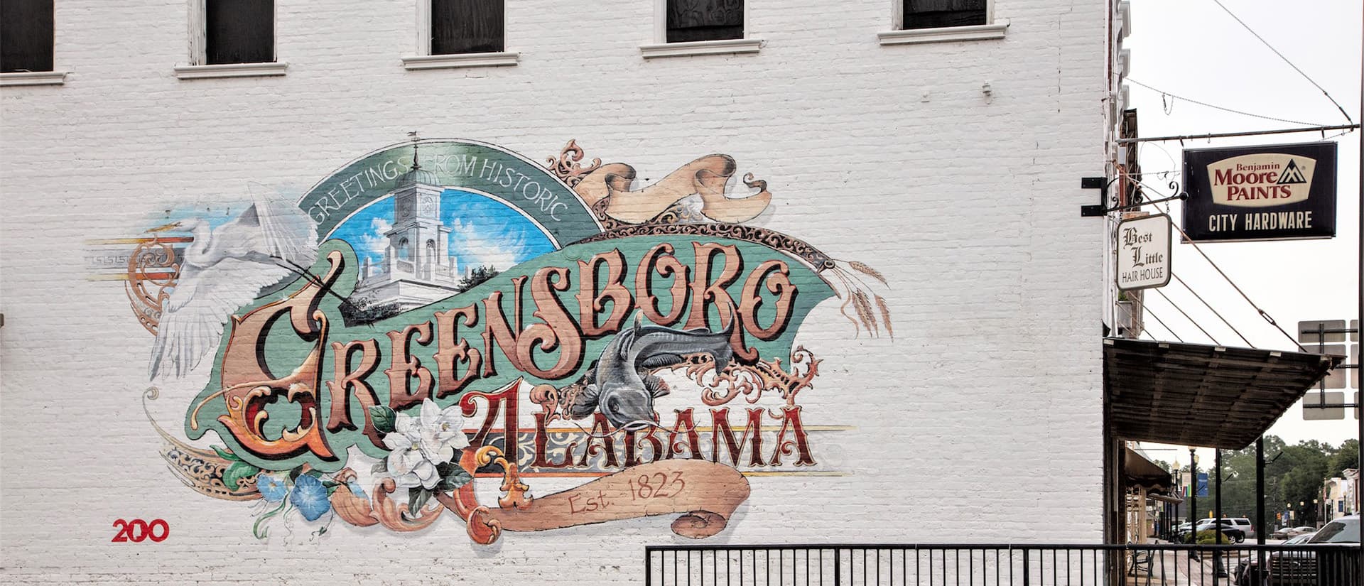 Mural depicting "Greensboro, Alabama" with historical elements on a brick wall.