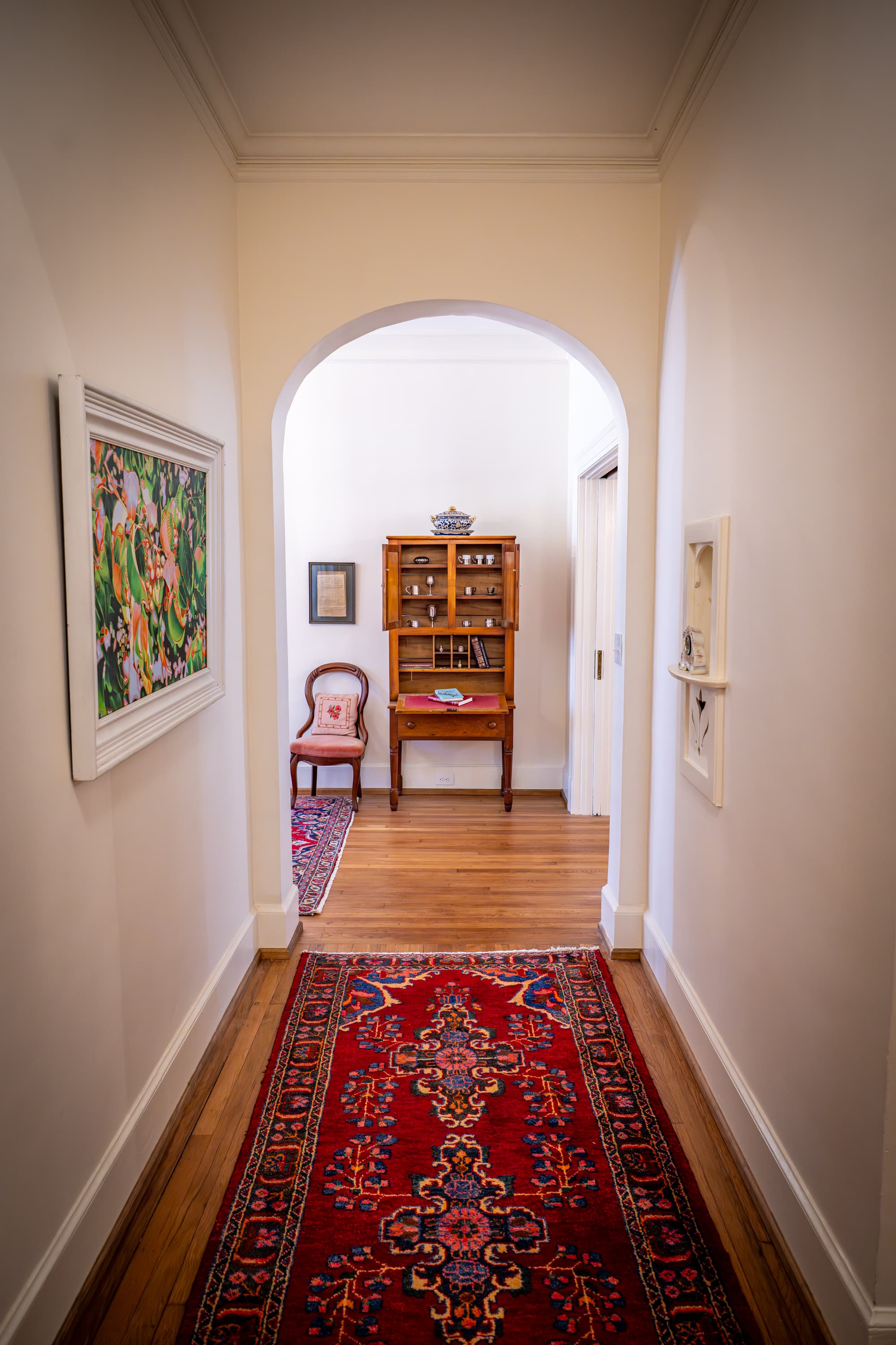 A view down a hallway with a patterned rug, framed art, and a small wooden cabinet at the end.
