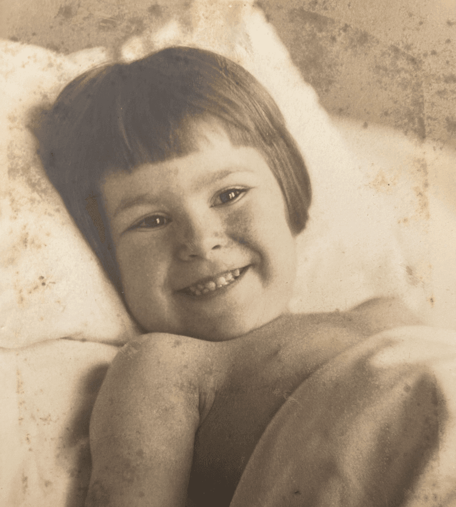 A smiling child with short hair is lying on a bed, partially covered by a blanket.