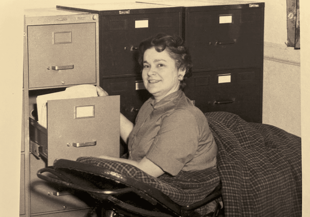 A woman smiles while sitting in a chair and organizing files in a cabinet.