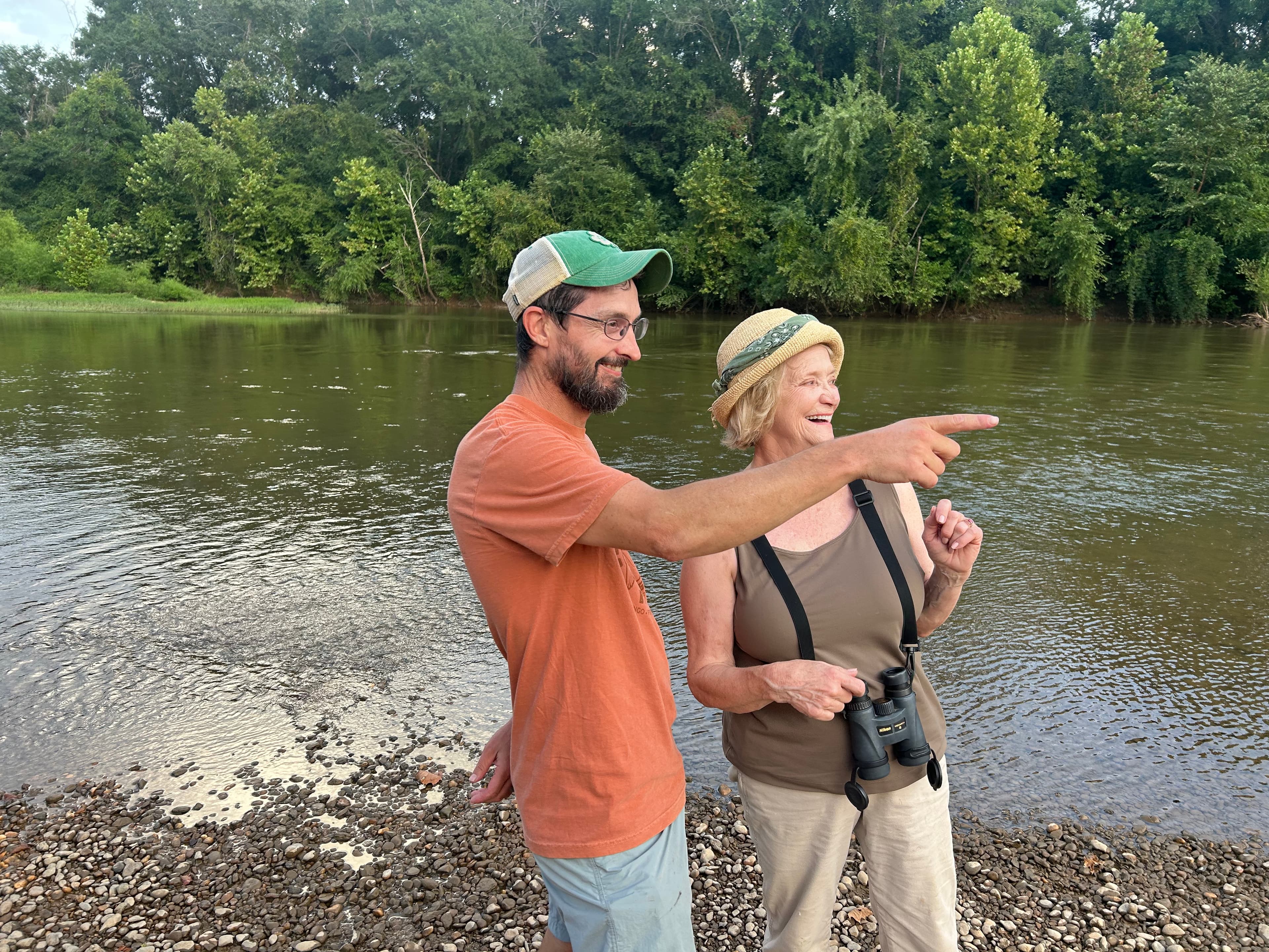 A man and a woman stand by a river, smiling and pointing at something in the distance.