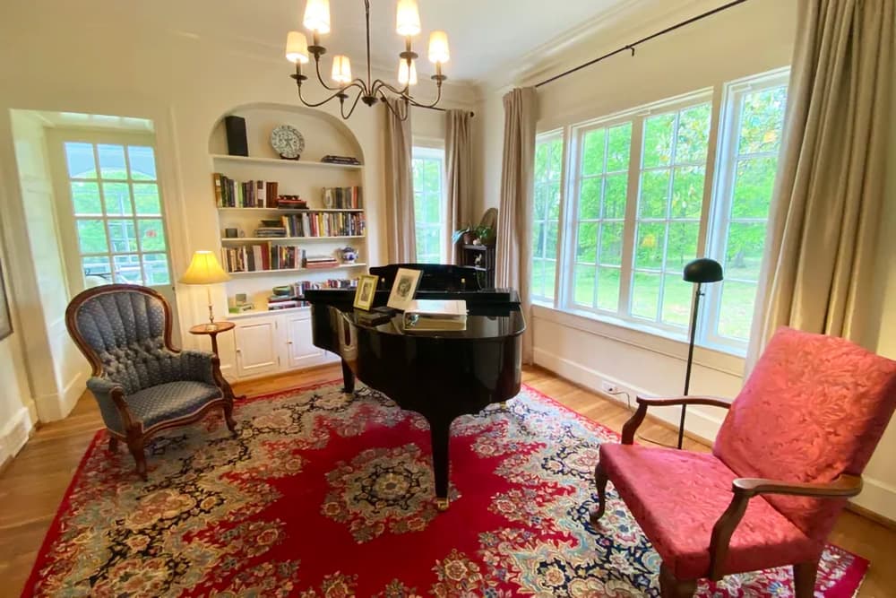 A cozy room featuring a grand piano, two armchairs, a bookshelf, and large windows overlooking greenery.