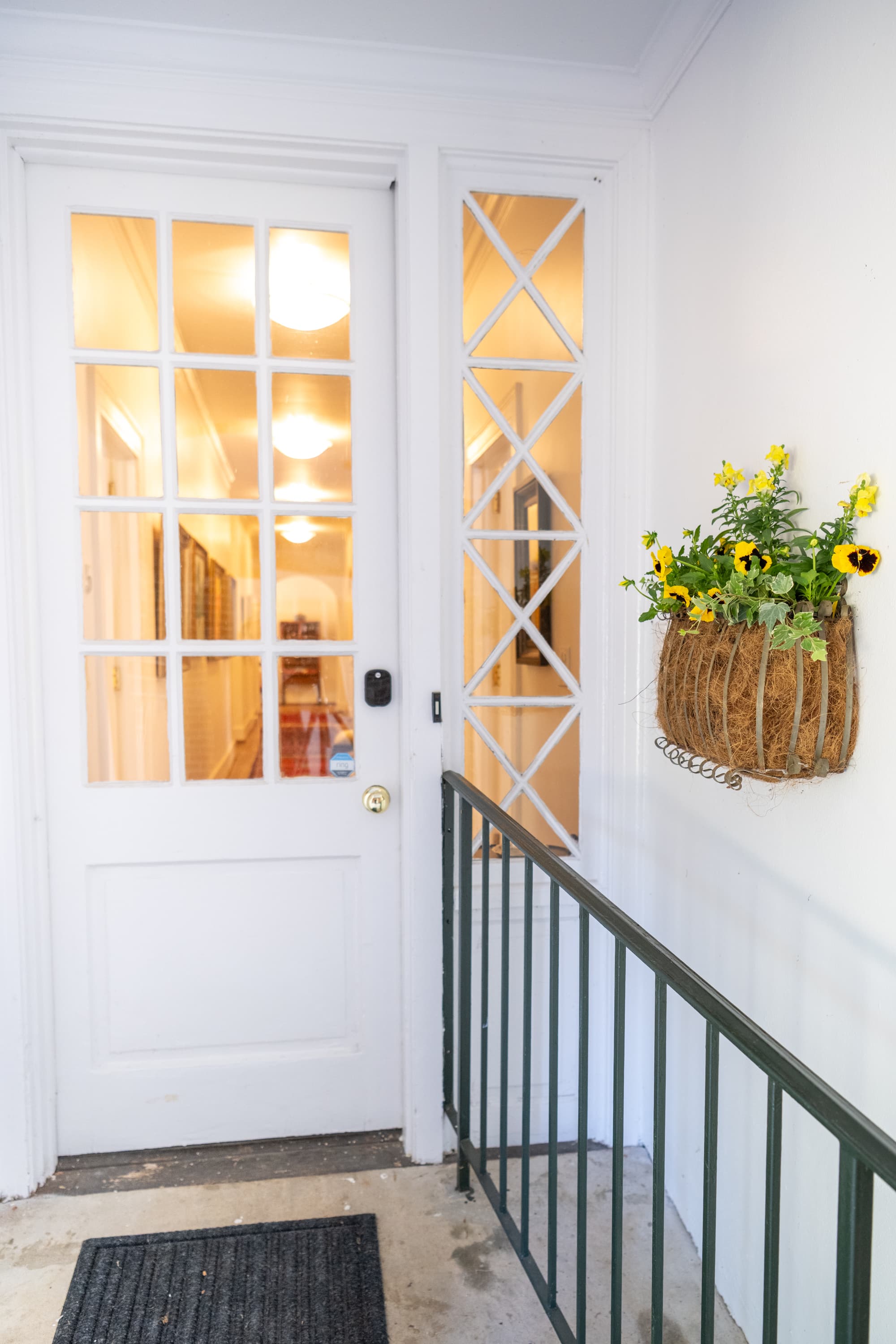 Welcoming white door featuring glass panes and a decorative X-pattern sidelight. A cheerful basket of yellow pansies hangs above the black railing, hinting at the warmth inside.