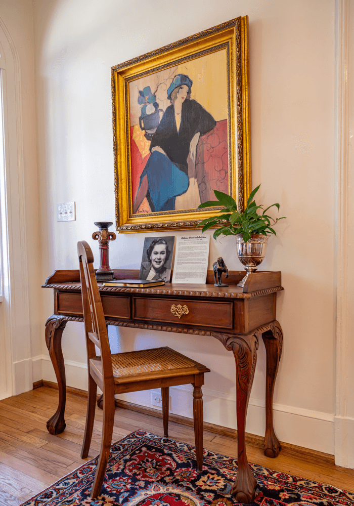 A vintage wooden desk with a chair, adorned with a plant, framed artwork, and a photo display on a colorful rug.