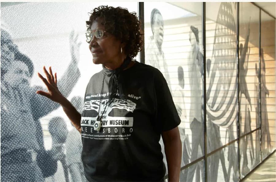 A woman in a Black History Museum t-shirt gestures towards a historical mural.