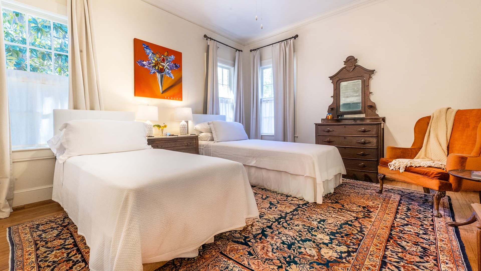 A cozy bedroom with two twin beds, an orange armchair, and a vintage dresser, illuminated by natural light.