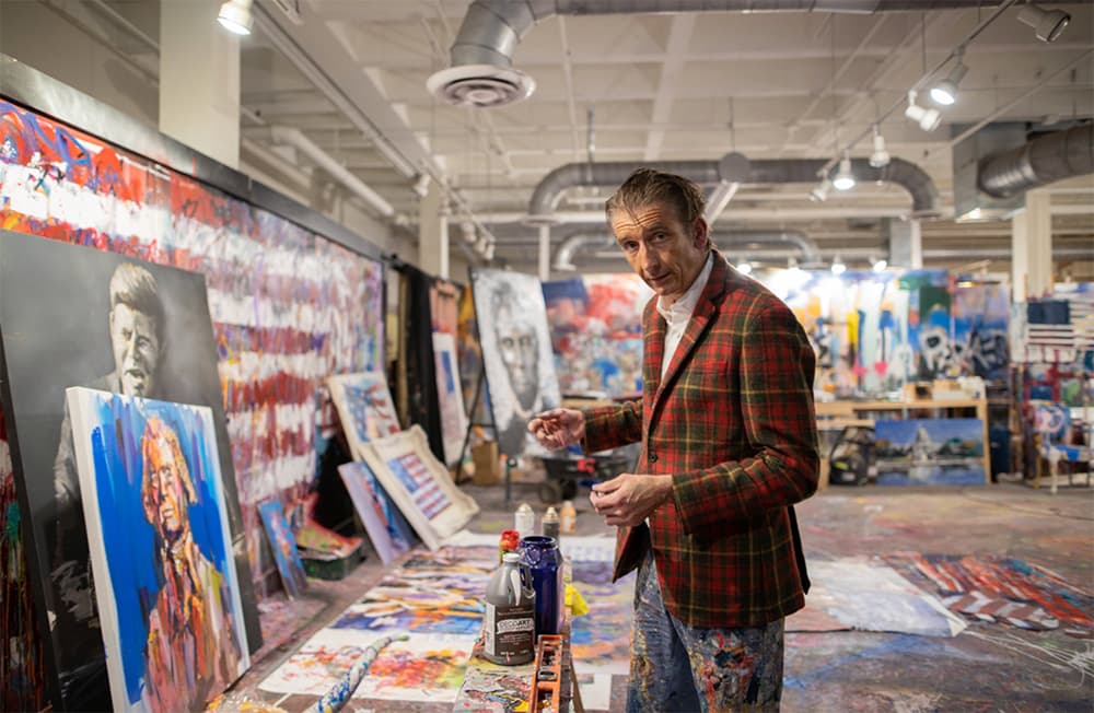 An artist in a plaid jacket stands in a colorful studio filled with various canvases and paint supplies.